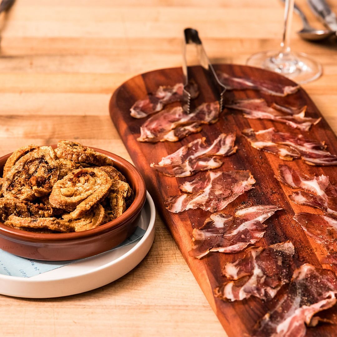 Close-up of tapas featuring crispy snacks in a bowl and cured meat slices on a wooden board, creating a rustic food presentation.