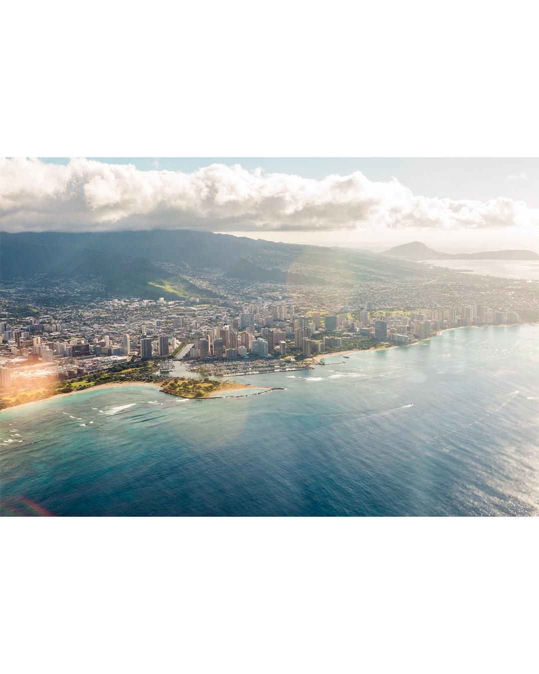 Aerial view of Honolulu cityscape, showing city buildings along the coastline, next to blue ocean and the island's mountain range.