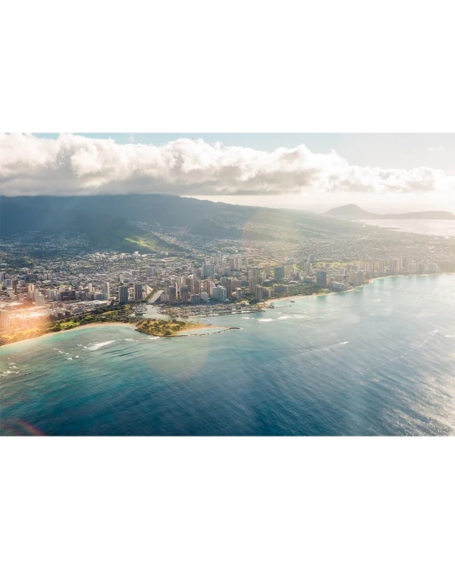 Aerial view of Honolulu cityscape, showing city buildings along the coastline, next to blue ocean and the island's mountain range.