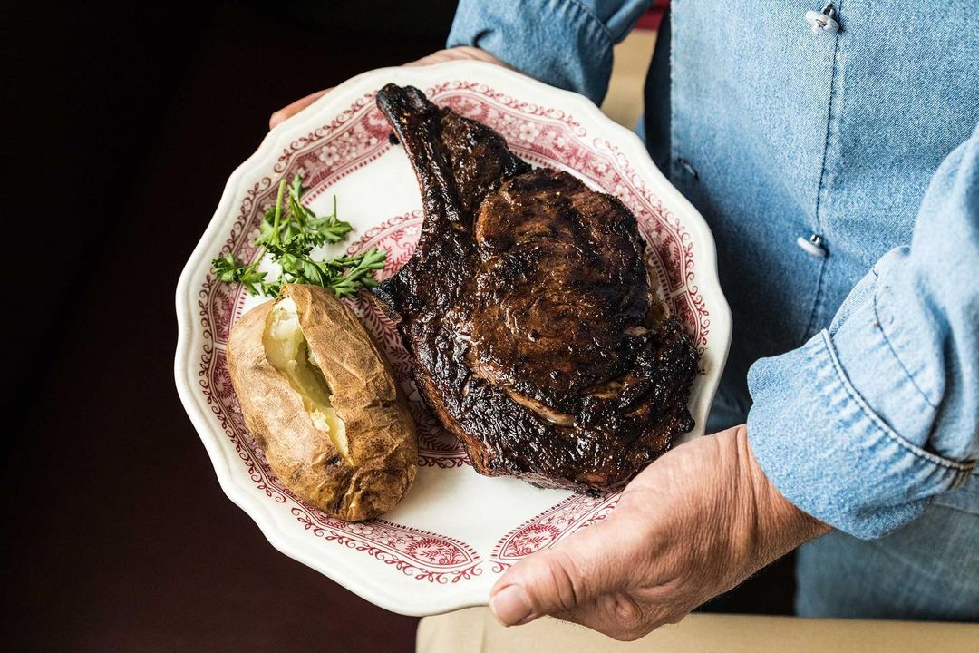 A person in a denim shirt holds a vintage plate with a cooked steak, baked potato, and herb garnish, presented in a close-up food shot.