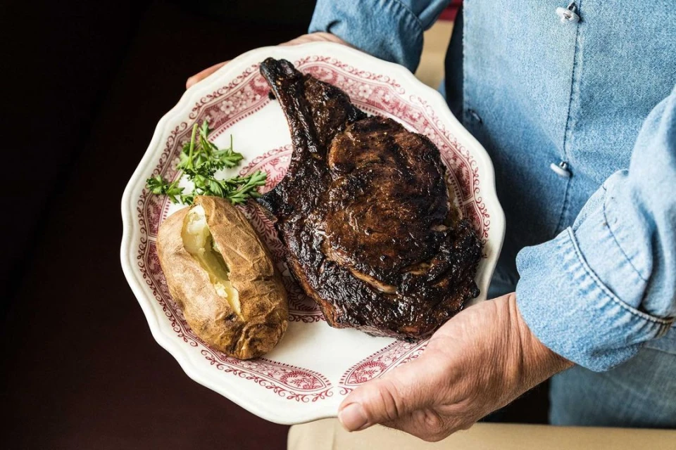 A person in a denim shirt holds a vintage plate with a cooked steak, baked potato, and herb garnish, presented in a close-up food shot.