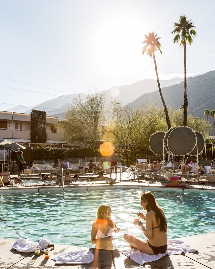Two women sit by a pool with other people lounging nearby, with palm trees and mountains visible under a bright, sunny sky.