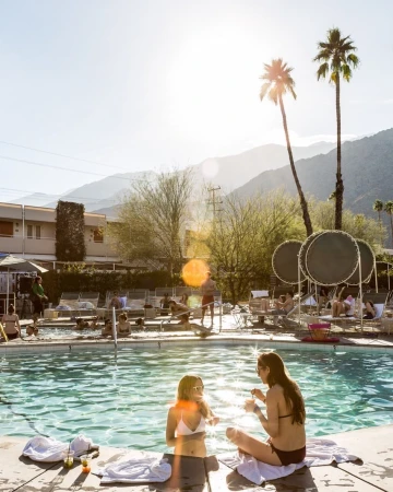 Two women sit by a pool with other people lounging nearby, with palm trees and mountains visible under a bright, sunny sky.