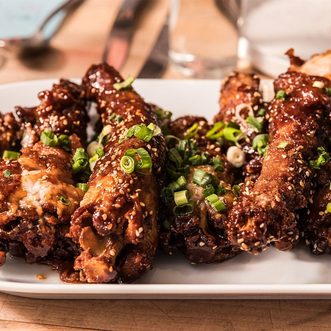 Close-up of crispy fried chicken wings glazed with a dark, glossy sauce, topped with sesame seeds and sliced scallions, on a white plate.