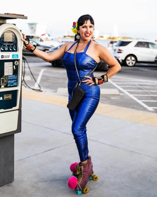 A woman with flowers in her hair and wearing a blue jumpsuit poses on roller skates next to a parking meter in a daytime outdoor shot.