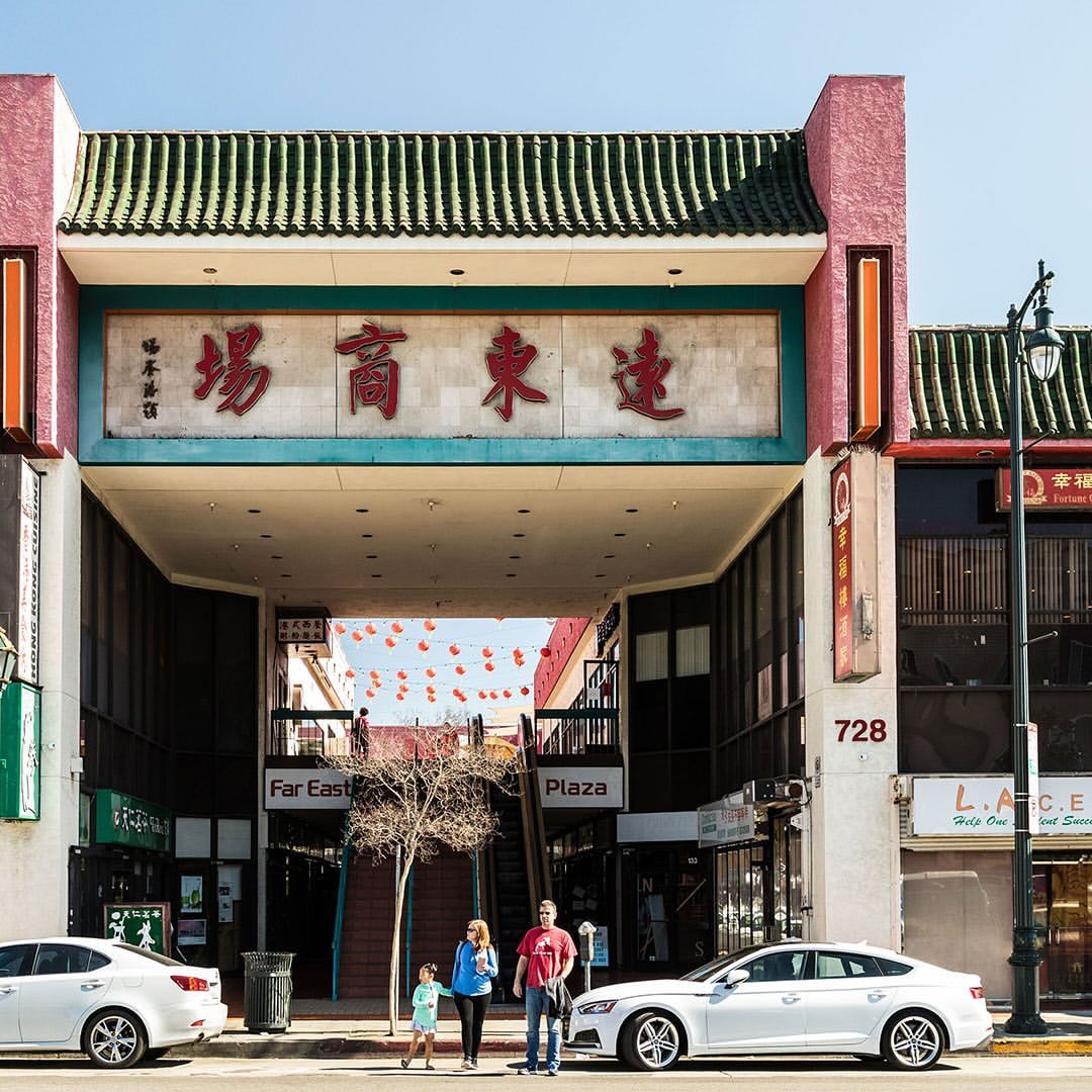 Family walks past Far East Plaza in Chinatown. Chinese characters adorn the plaza's entrance; two cars are parked on the street.