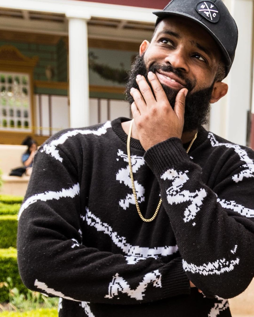 Portrait of a bearded man wearing a patterned sweater and ball cap, set in an outdoor courtyard with white pillars.