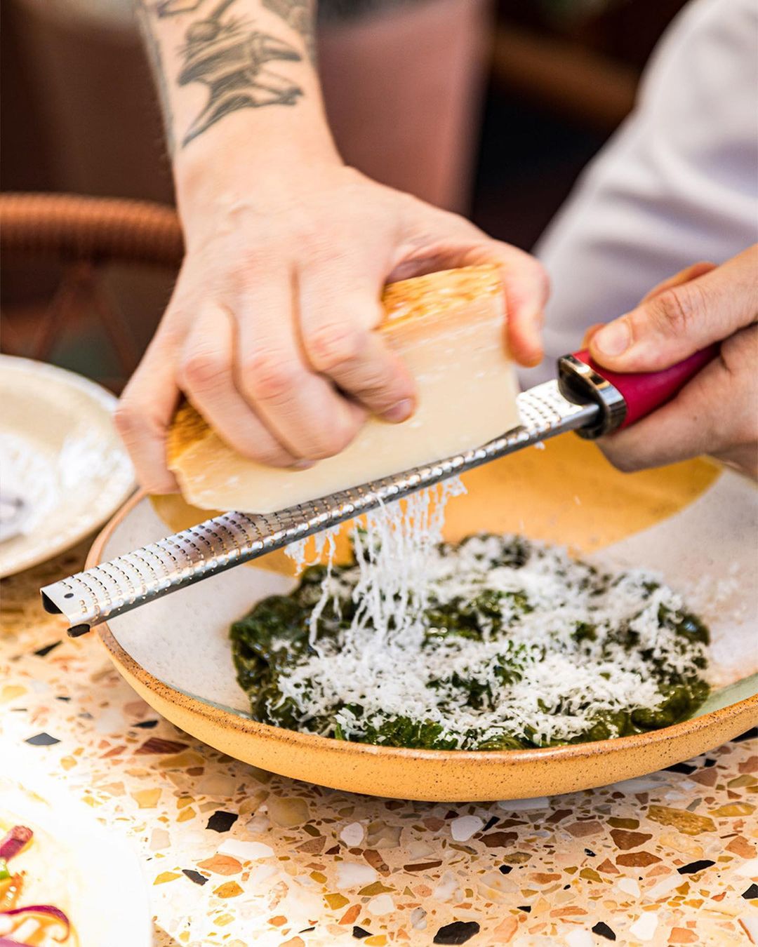 Tattooed adult hands grating parmesan cheese over a green vegetable dish on a speckled table, shot in a brightly lit indoor setting.