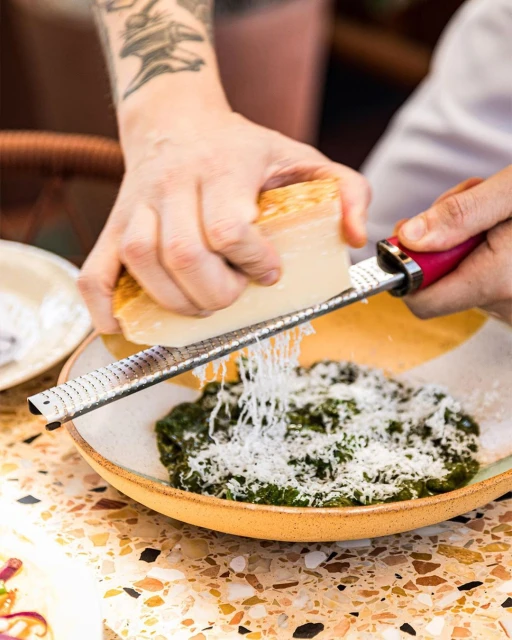Tattooed adult hands grating parmesan cheese over a green vegetable dish on a speckled table, shot in a brightly lit indoor setting.