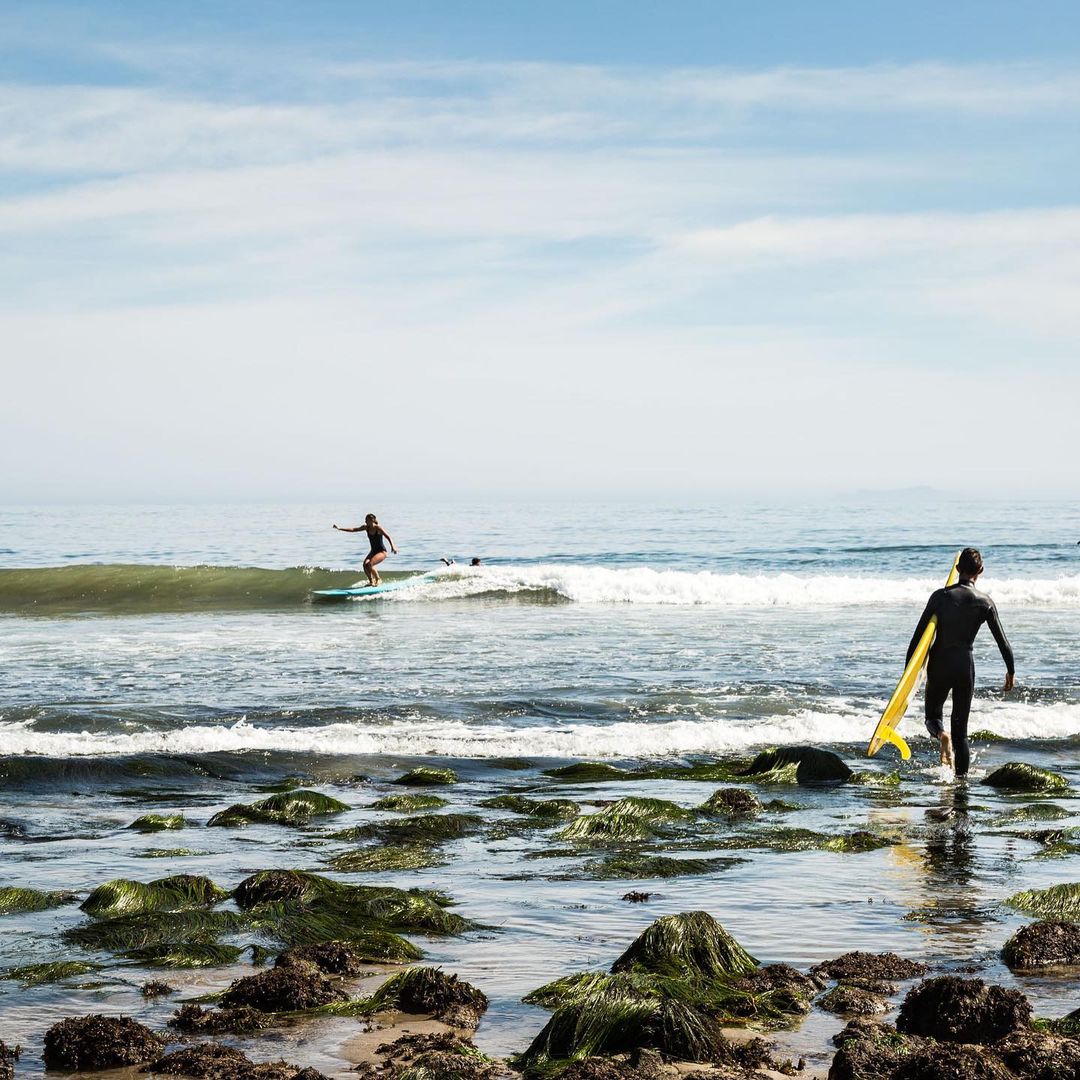Two surfers, one riding a wave, the other carrying a yellow surfboard while walking out of the water onto a rocky beach.