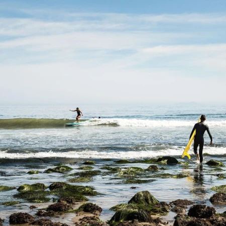 Two surfers, one riding a wave, the other carrying a yellow surfboard while walking out of the water onto a rocky beach.