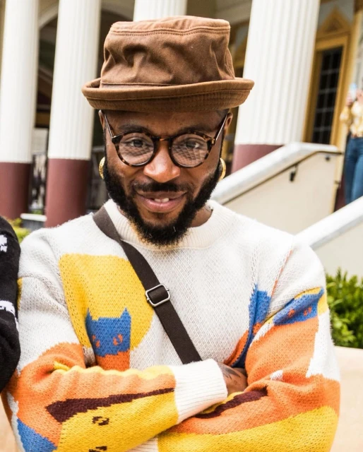 A smiling black man with glasses and a bucket hat poses outdoors wearing a patterned sweater and crossbody bag in a medium studio shot.