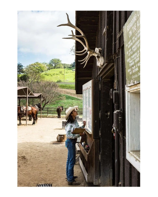 Woman in cowboy hat checks a board outside a wooden building on a ranch with horses and a green field under a partly cloudy sky.