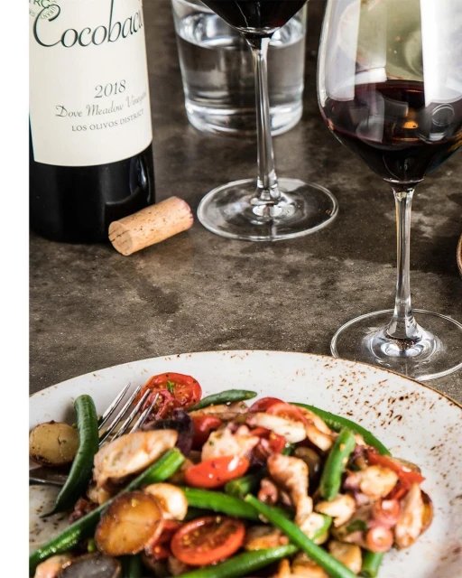 Overhead shot of a wine bottle, glasses of red wine, and a plate of vegetables with protein on a dark concrete surface.