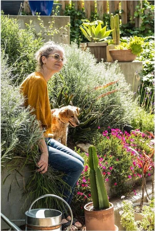 Woman and dog together in a lush garden with concrete planters, flowers, succulents, and a metal watering can on a sunny day.