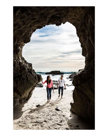 Man and woman walking on a sandy beach viewed through a large natural rock cave opening