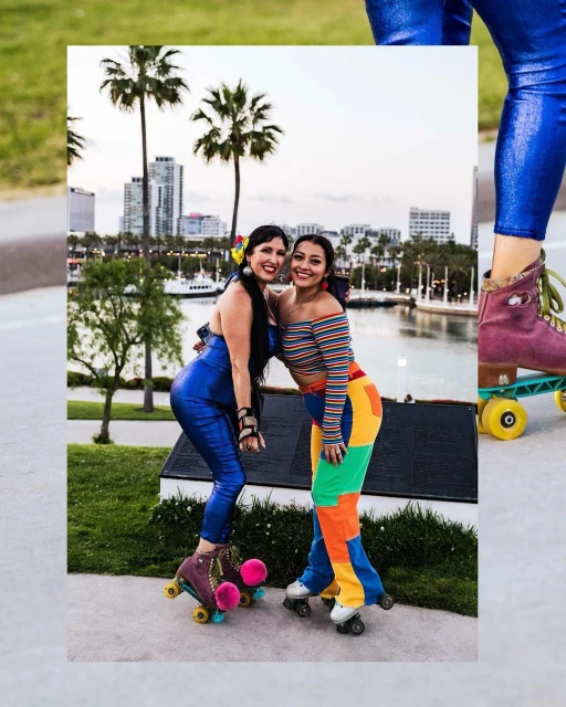 Two adult women pose side-by-side in colorful vintage clothing and roller skates with an urban park and cityscape in the background.