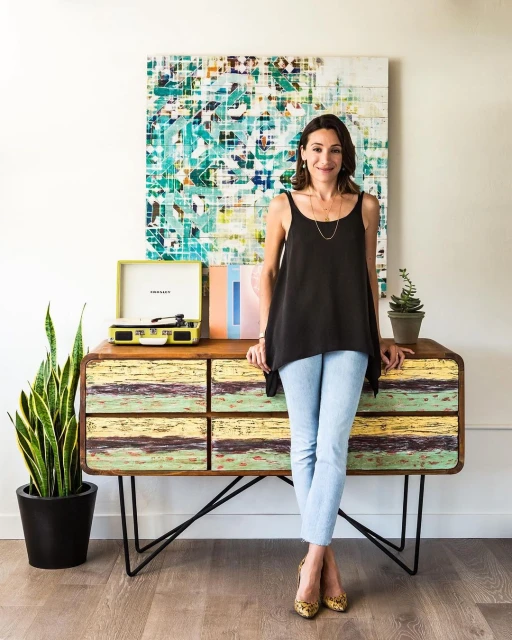 Woman standing next to a console table in an indoor environment with a potted plant and abstract art piece in the background.