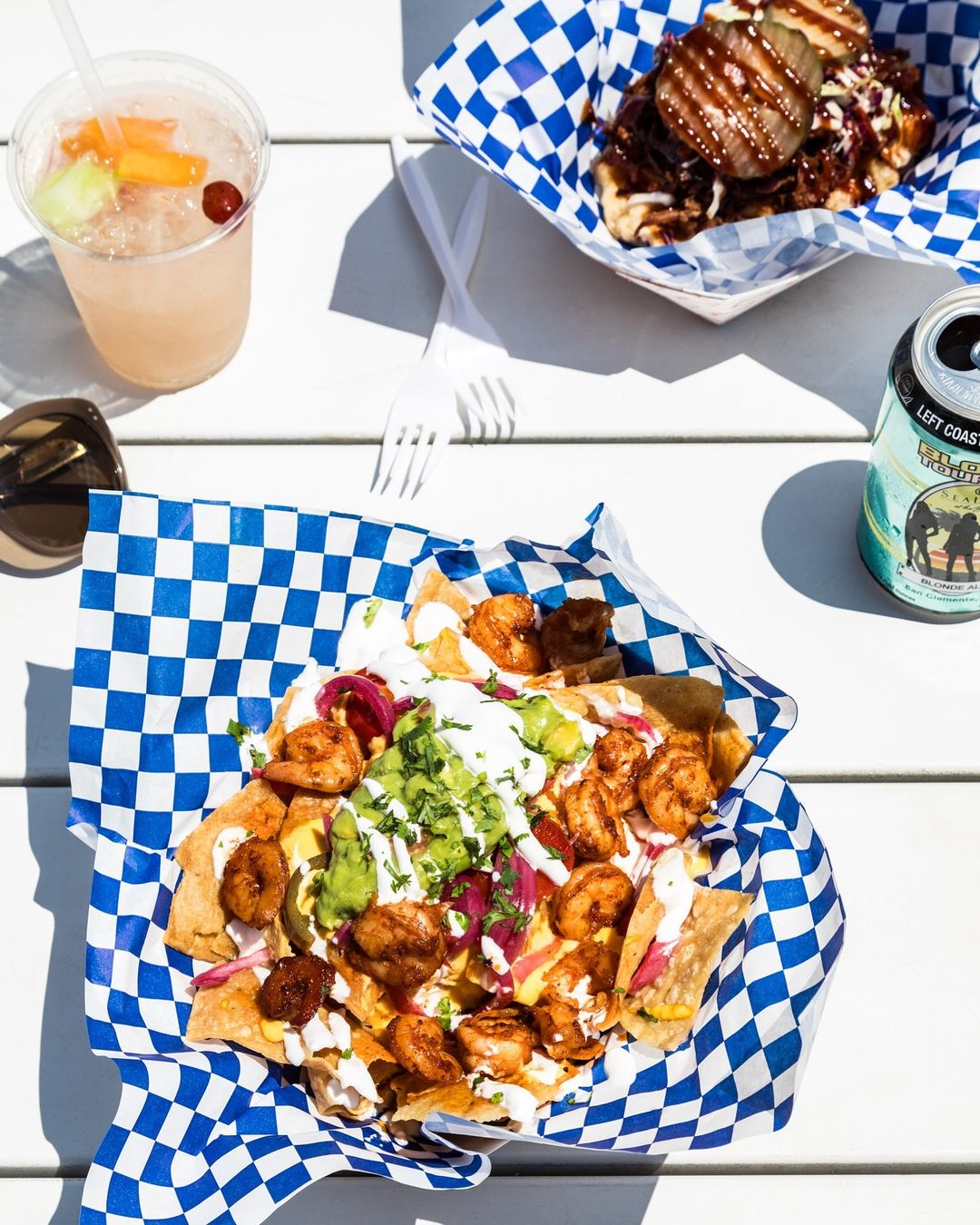 Overhead shot of shrimp nachos, BBQ waffle fries, and a cocktail on a white table with bright sunlight and blue checkered liners.