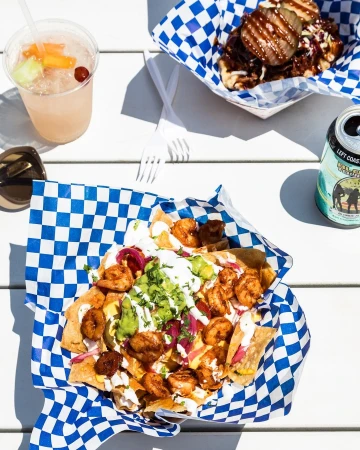 Overhead shot of shrimp nachos, BBQ waffle fries, and a cocktail on a white table with bright sunlight and blue checkered liners.
