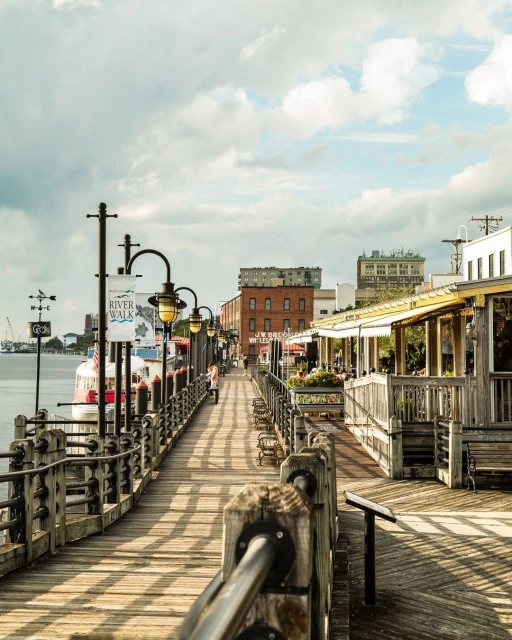 Scenic view of River Walk in Savannah, Georgia, showcasing a charming boardwalk lined with benches, lamp posts, and historic buildings.