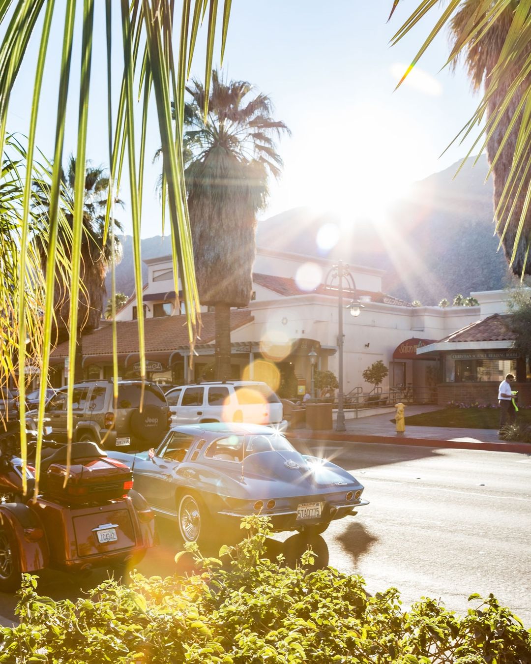 Palm Springs street view with vintage blue car and motorcycle parked near palm trees, businesses, and distant mountains on a sunny day.