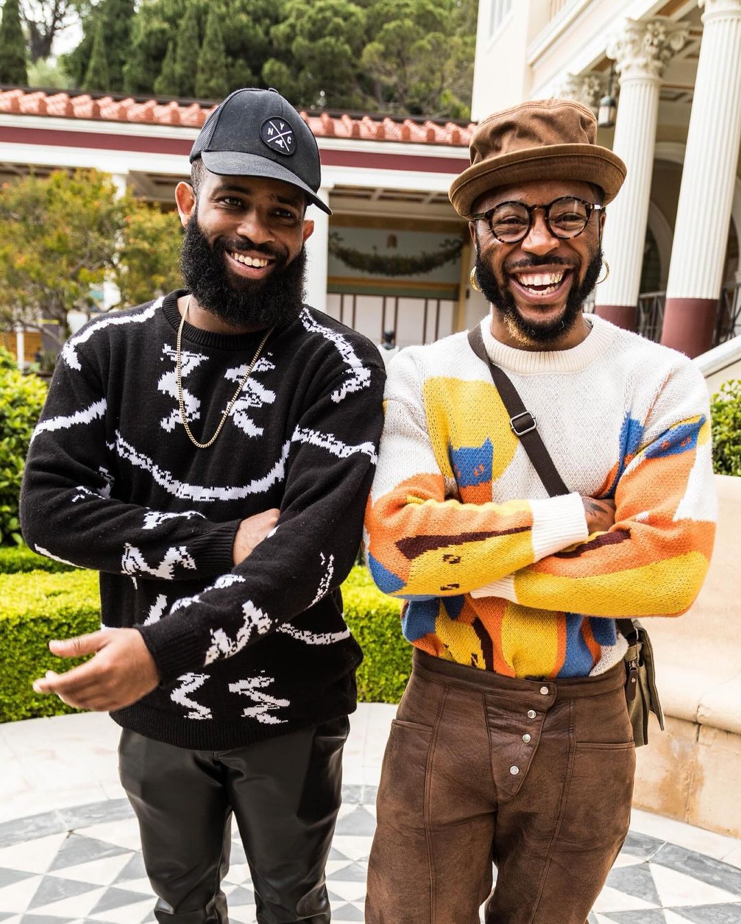 Two bearded men pose in front of a mansion with columns, wearing hats and patterned sweaters. One man wears leather pants.