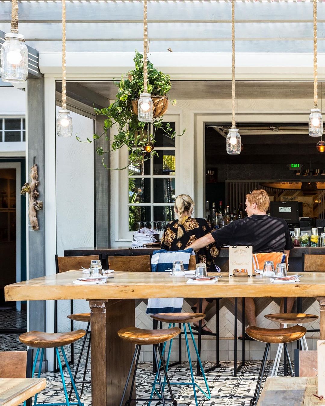 Outdoor restaurant scene with a couple sitting at a bar, illuminated by hanging pendant lights and natural light, creating a warm atmosphere.