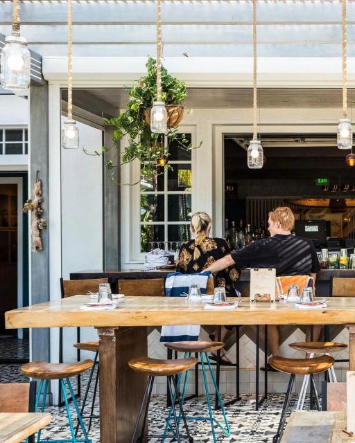 Outdoor restaurant scene with a couple sitting at a bar, illuminated by hanging pendant lights and natural light, creating a warm atmosphere.