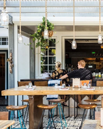 Outdoor restaurant scene with a couple sitting at a bar, illuminated by hanging pendant lights and natural light, creating a warm atmosphere.