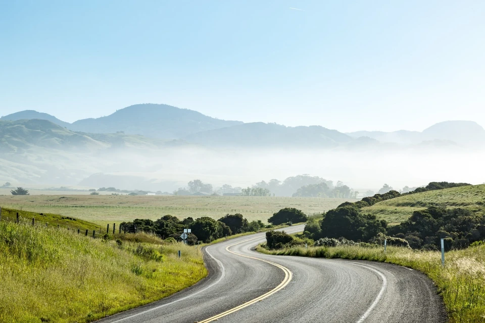 Winding asphalt road curves through green grassy hillsides with fog-shrouded mountains in the background under a blue sky.