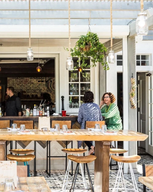 Outdoor restaurant scene with two women seated at a wooden table, glasses, bar with bottles, hanging mason jar lights, and plants.