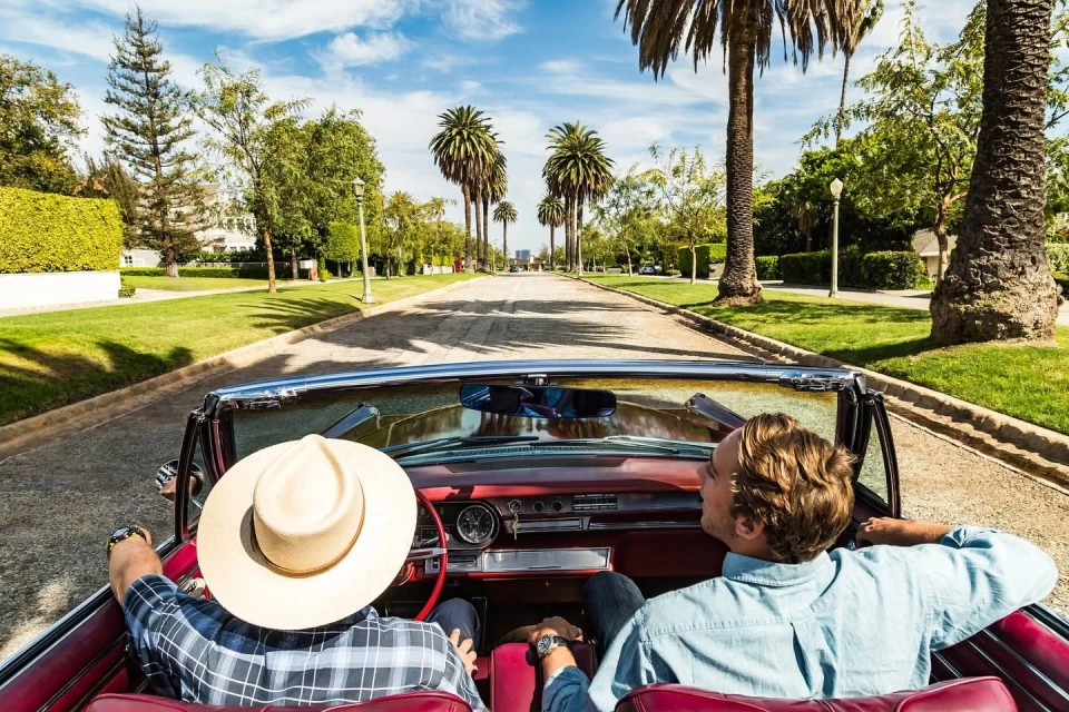 Two adult men drive a red convertible on a road lined with green lawns and palm trees under a blue sky with some clouds.