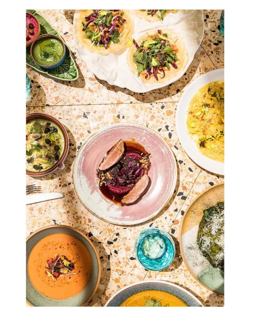 Overhead shot of colorful plates of food, featuring tacos, soups, and vegetable dishes, arranged on a terrazzo table.