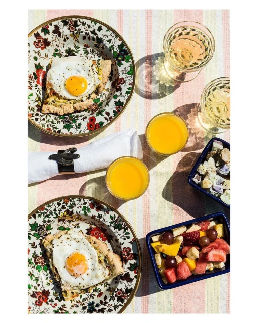 Overhead shot of breakfast spread: sunny-side-up eggs on floral plates, orange juice, fruit salad, white wine on striped tablecloth.