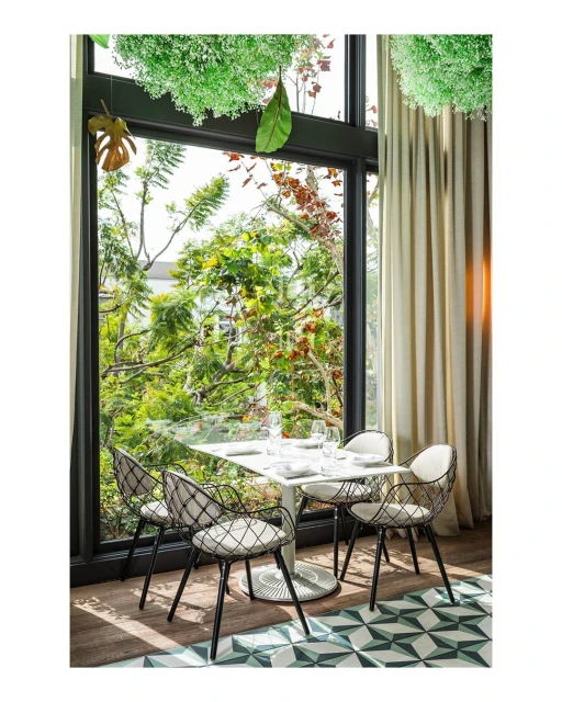 Indoor restaurant setting with a square table and four modern chairs in front of a large window with greenery outside and hanging decor.