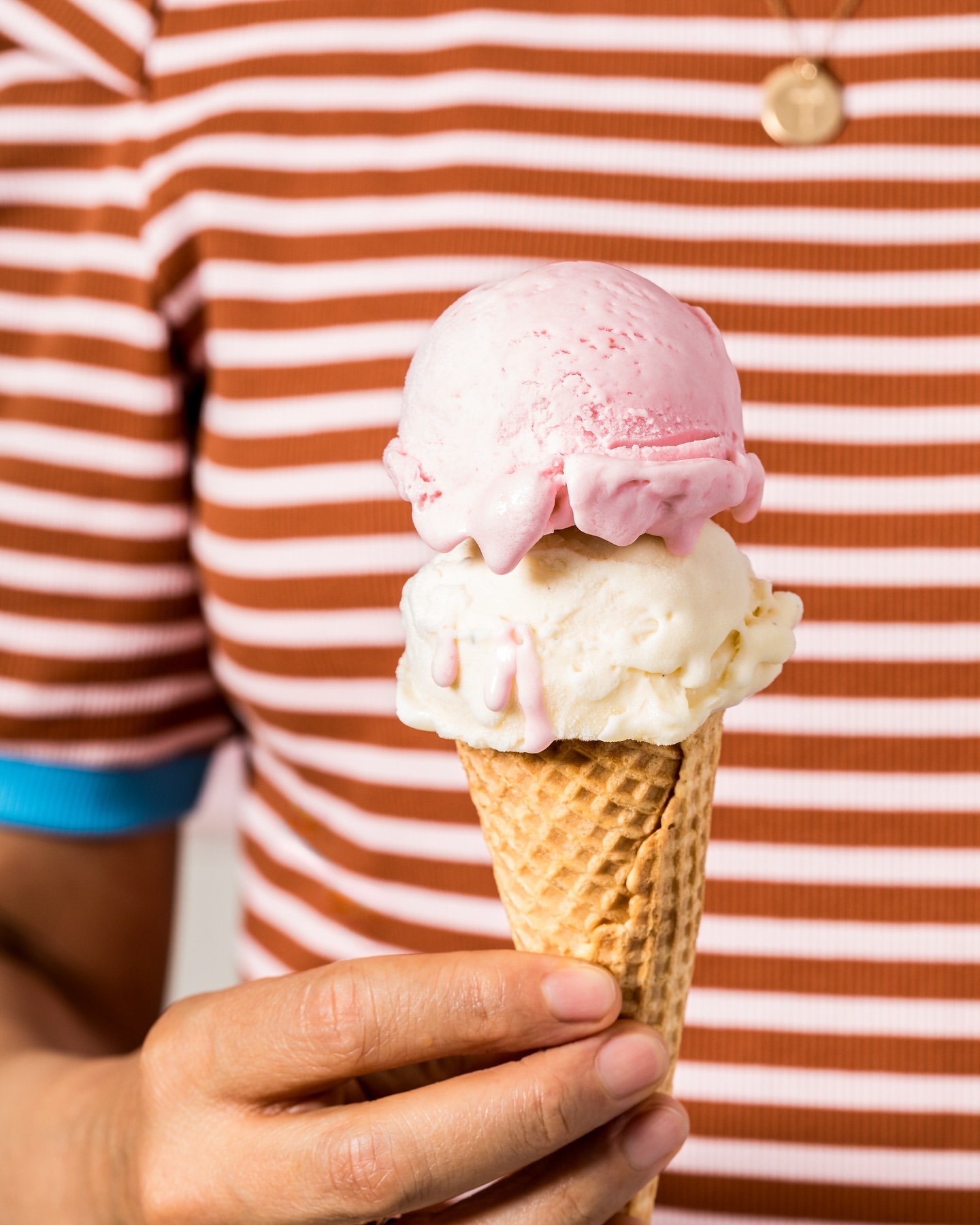 Person holding a waffle cone with two scoops of ice cream, pink and vanilla, against a striped brown and white background.