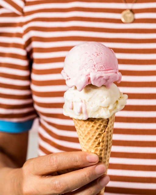 Person holding a waffle cone with two scoops of ice cream, pink and vanilla, against a striped brown and white background.