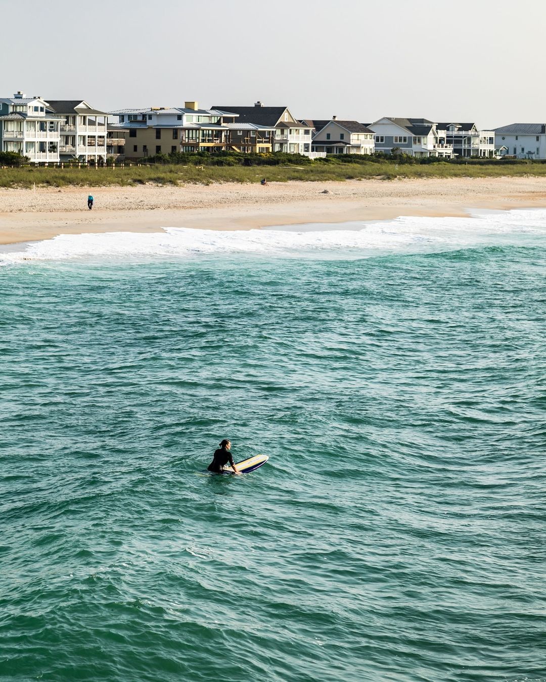Surfer sitting on a surfboard in the ocean water near a sandy beach and residential houses on a sunny day, creating a serene coastal scene.