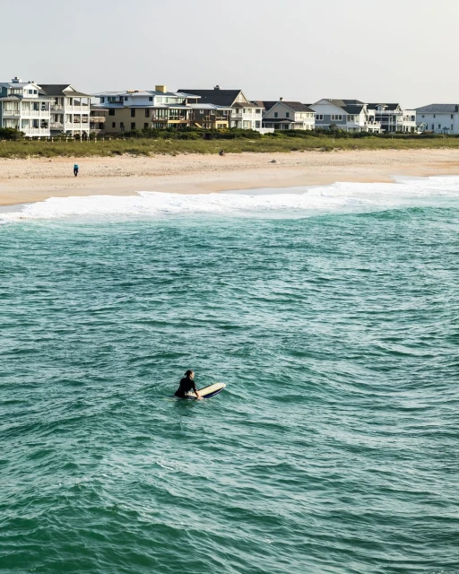 Surfer sitting on a surfboard in the ocean water near a sandy beach and residential houses on a sunny day, creating a serene coastal scene.
