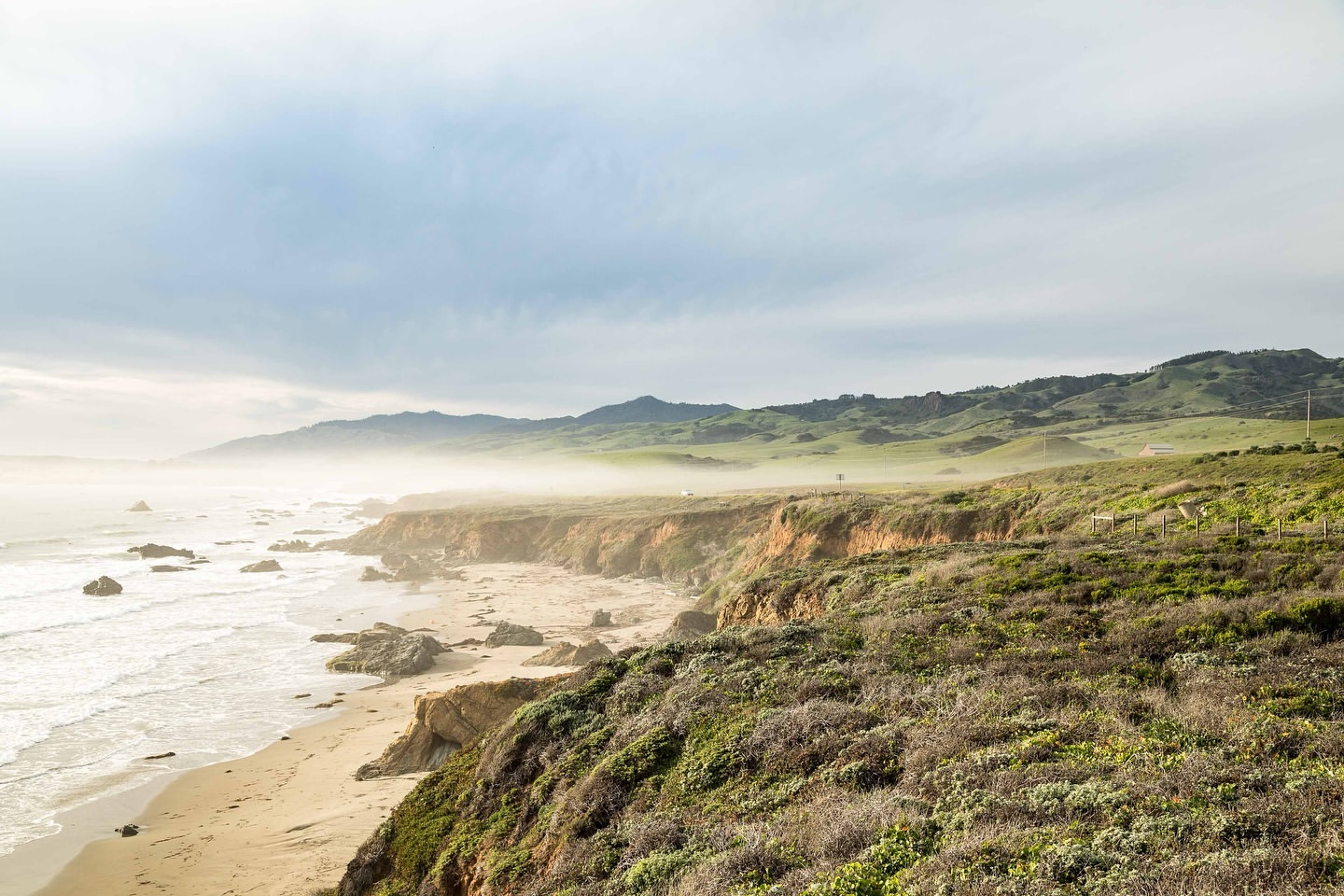 Coastal landscape featuring a rocky shoreline, sandy beach, and green hills under an overcast sky, capturing a serene seascape.