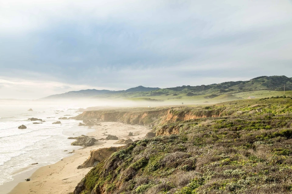 Coastal landscape featuring a rocky shoreline, sandy beach, and green hills under an overcast sky, capturing a serene seascape.