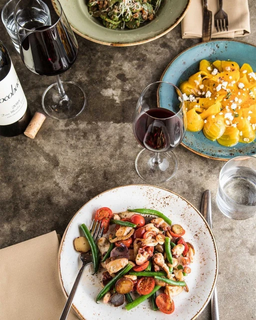 Overhead shot of culinary scene with plates of colorful salads and pasta, glasses of red wine, and silverware on a neutral table.