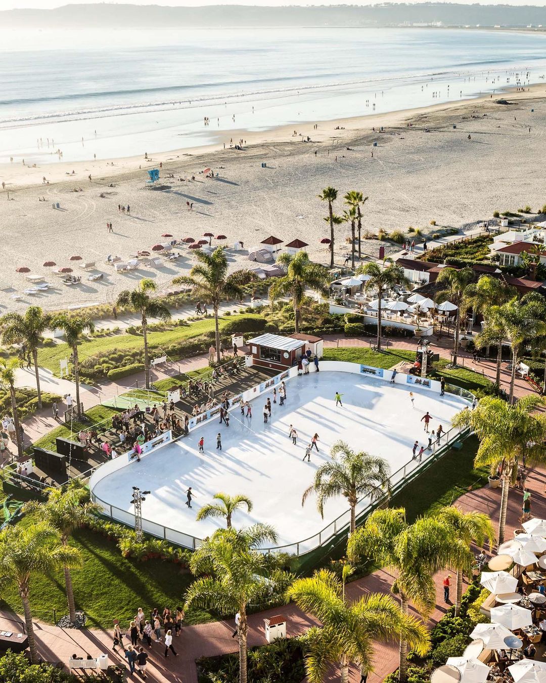 Scenic aerial view of an outdoor ice skating rink surrounded by palm trees, with a beach and ocean in the background, bathed in sunlight.