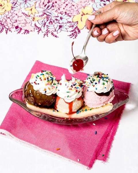 Banana split in a glass dish being topped with a cherry from a spoon, set on a pink napkin with a floral tablecloth background.