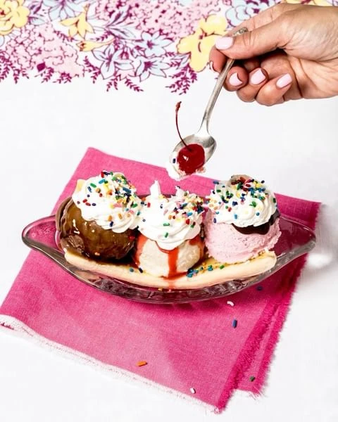 Banana split in a glass dish being topped with a cherry from a spoon, set on a pink napkin with a floral tablecloth background.