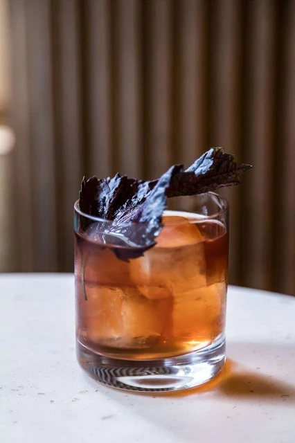 A dark cocktail in a clear rocks glass with a large ice cube, garnished with dark green leaves on a white marble table.