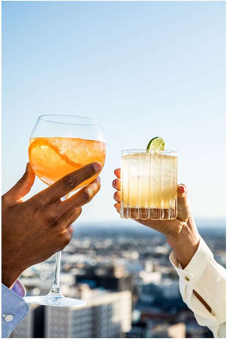 Two cocktails are held up in celebration with a city skyline and bright blue sky in the background, creating a celebratory toast.
