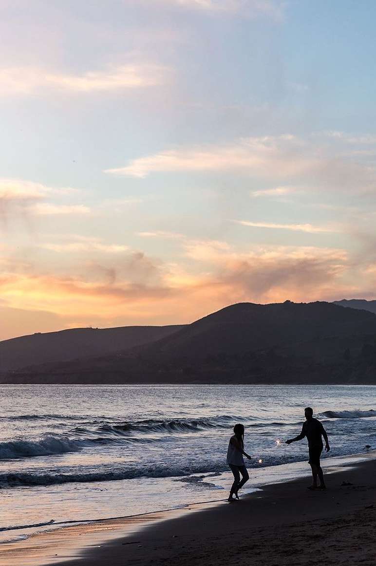 Silhouetted figures holding sparklers on a sandy beach at sunset with rolling hills in the background.