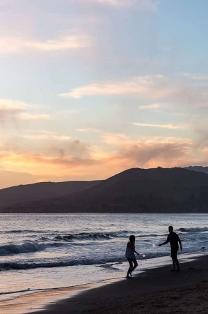 Silhouetted figures holding sparklers on a sandy beach at sunset with rolling hills in the background.
